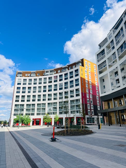 Exterior view of a modern residential building with a curved facade, featuring white walls accented by sections of orange, pink, and beige panels, under a bright blue sky with scattered clouds. The gr
